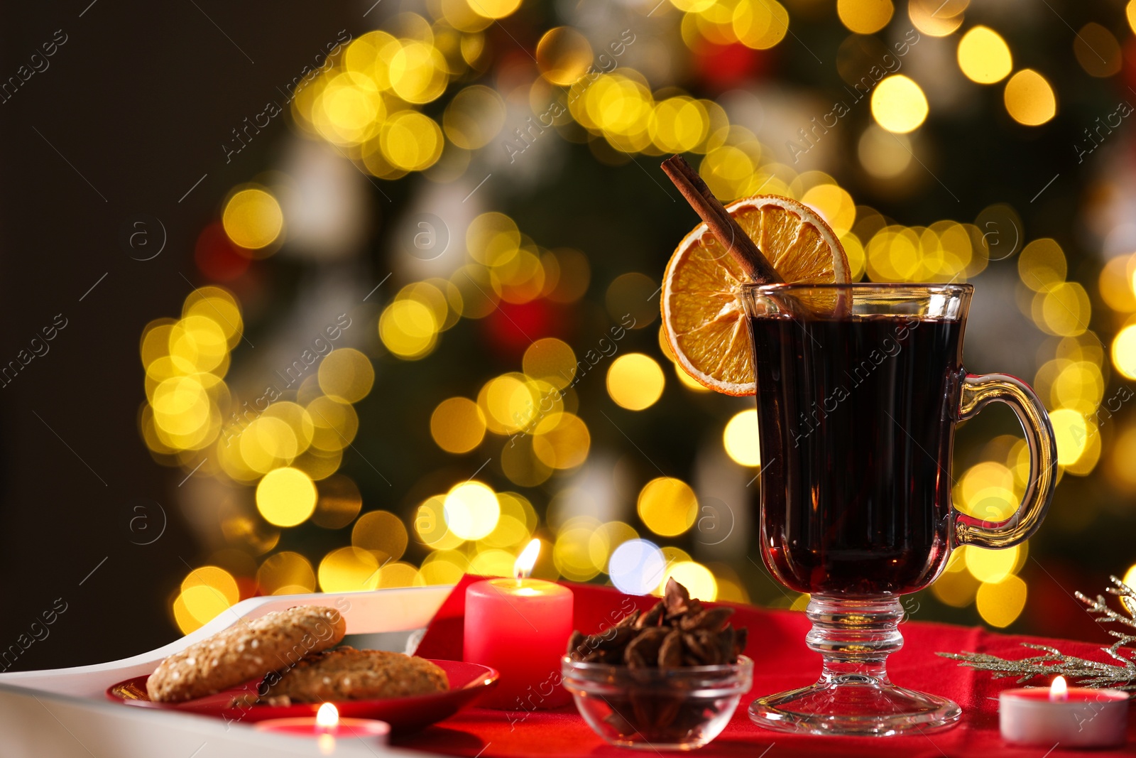 Photo of Aromatic mulled wine in glass cup, anise, cookies and burning candles on tray against blurred Christmas lights, bokeh effect