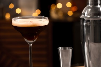 Photo of Delicious espresso martini in glass, jigger and shaker against blurred lights, closeup