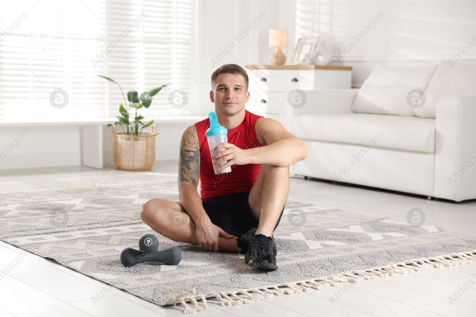 Photo of Athletic man with shaker of protein drink sitting on carpet at home