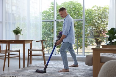 Photo of Young man cleaning rug with cordless vacuum cleaner at home