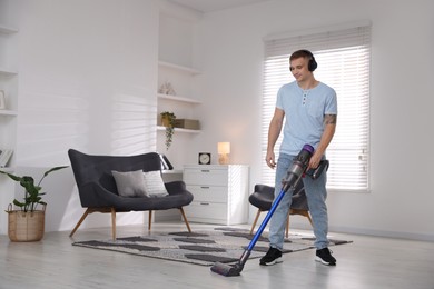 Photo of Young man in headphones cleaning floor with cordless vacuum cleaner at home