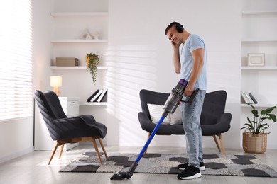 Photo of Young man in headphones cleaning floor with cordless vacuum cleaner at home