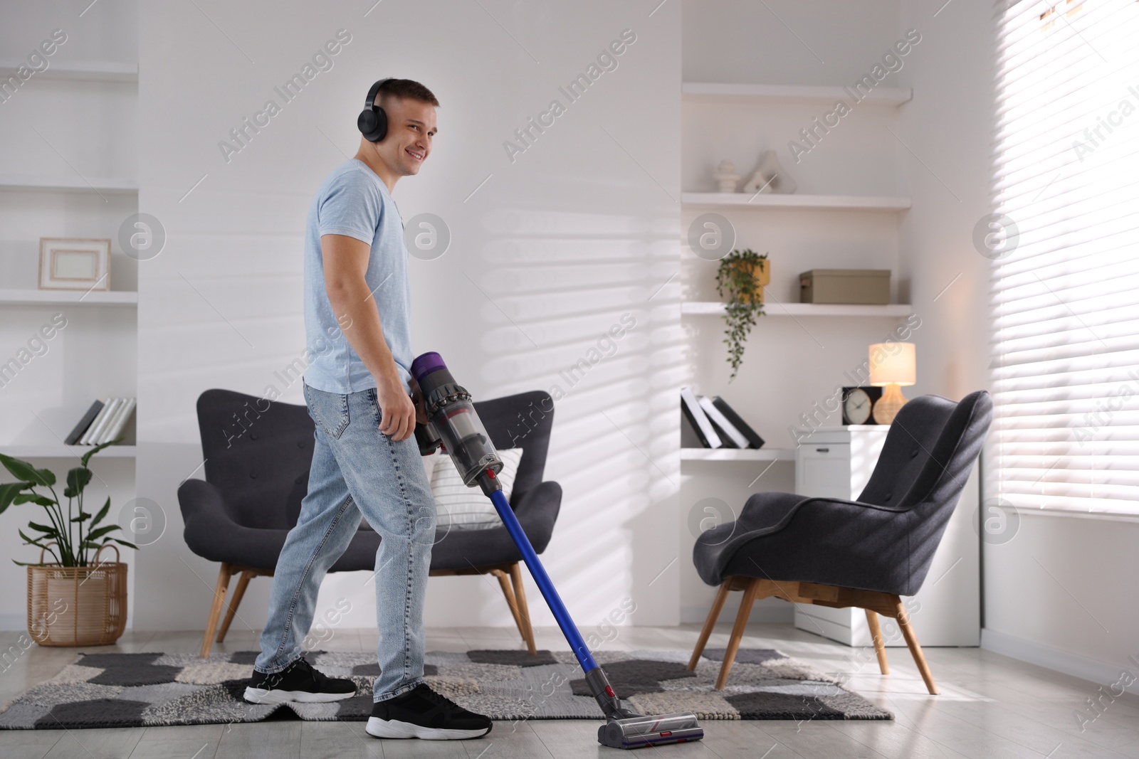 Photo of Young man in headphones cleaning floor with cordless vacuum cleaner at home