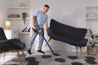 Photo of Young man cleaning rug under sofa with cordless vacuum cleaner at home