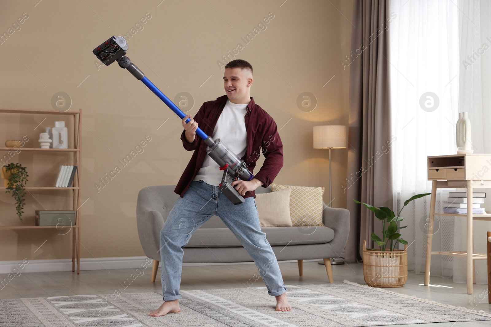 Photo of Young man having fun while cleaning rug with cordless vacuum cleaner at home