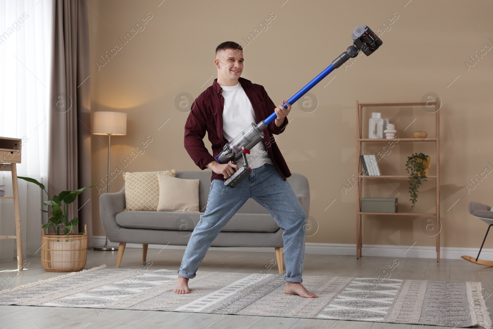 Photo of Young man having fun while cleaning rug with cordless vacuum cleaner at home