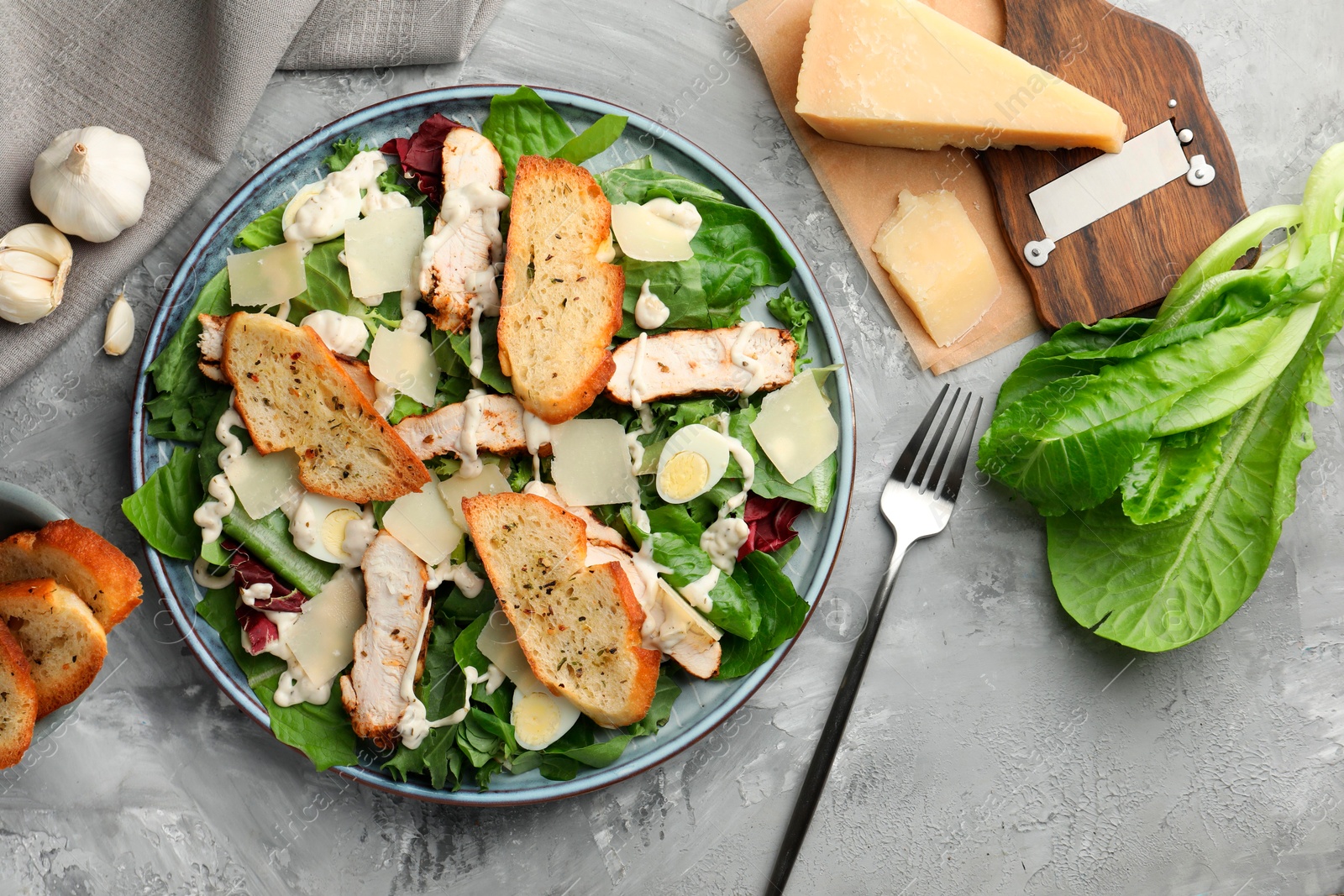 Photo of Tasty Caesar salad with chicken, ingredients and fork on gray textured table, flat lay