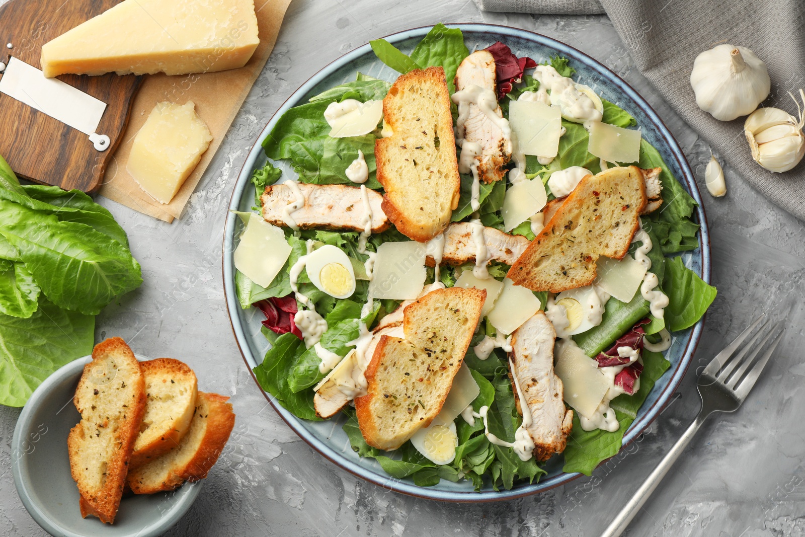 Photo of Tasty Caesar salad with chicken, ingredients and fork on gray textured table, flat lay