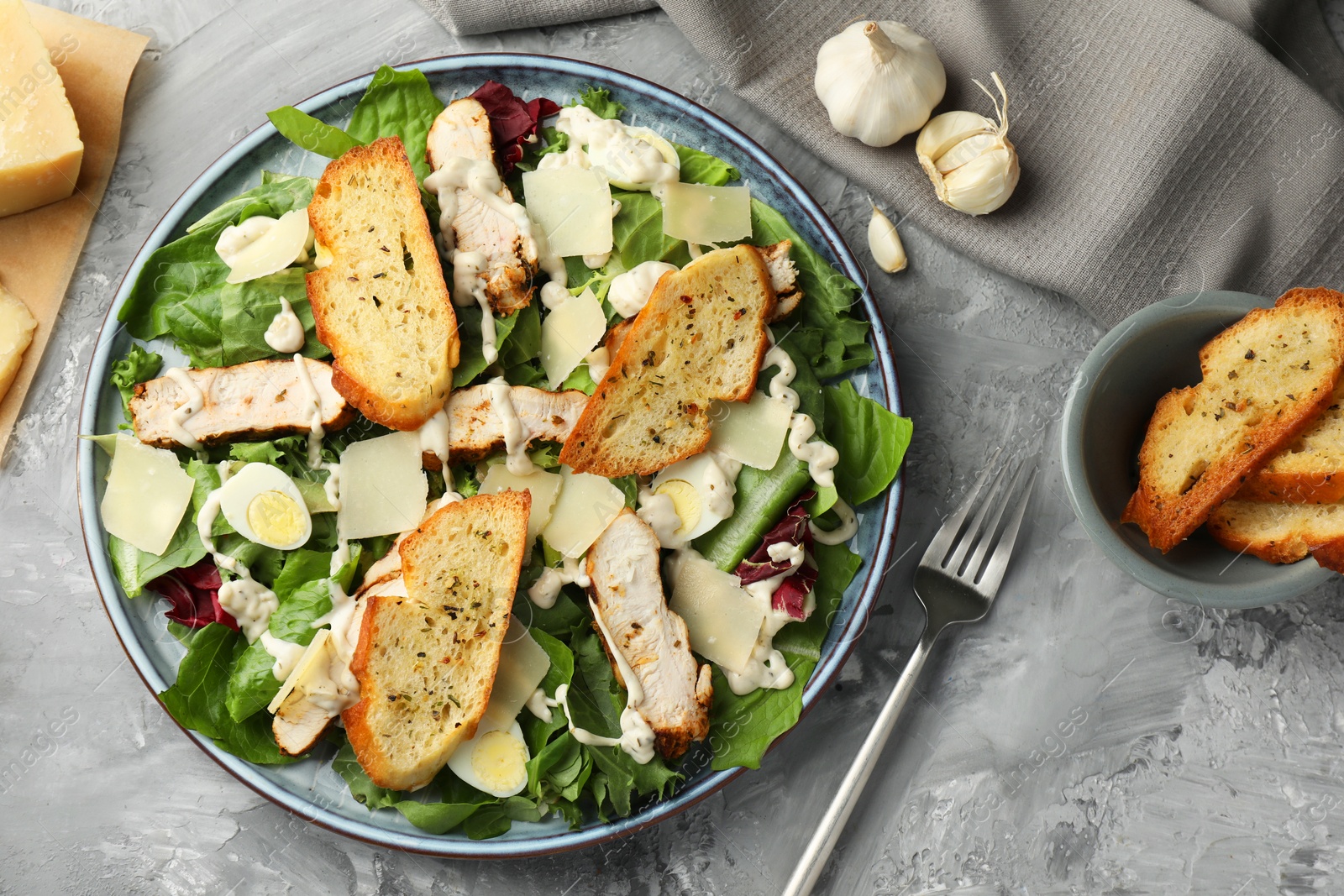 Photo of Tasty Caesar salad with chicken, ingredients and fork on gray textured table, flat lay