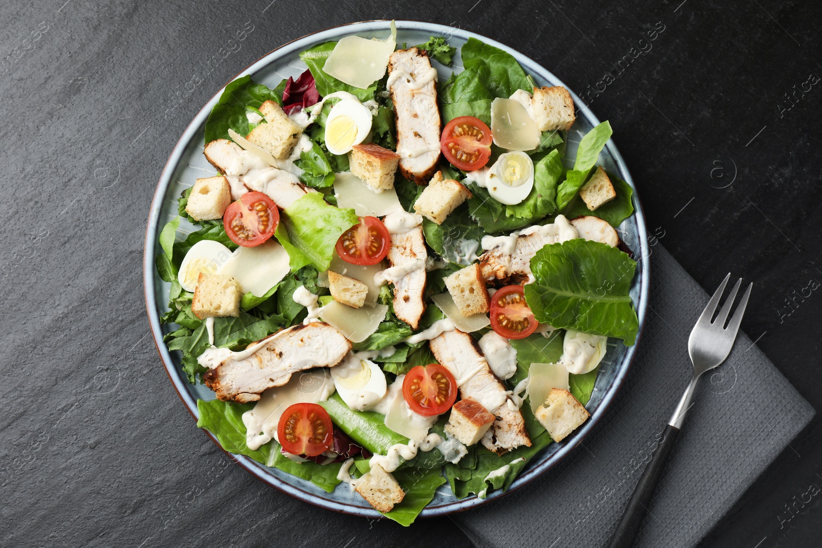 Photo of Tasty Caesar salad with chicken and tomatoes served on dark gray textured table, flat lay