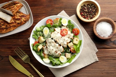 Photo of Tasty Caesar salad with chicken and tomatoes in bowl served on wooden table, flat lay