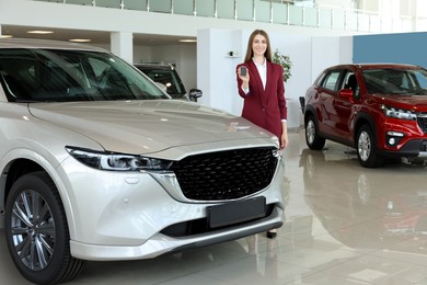 Photo of Happy saleswoman holding key near new silver car in salon