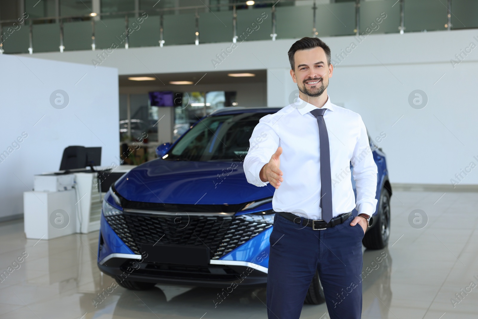 Photo of Happy salesman reaching for handshake with client near new blue car in salon