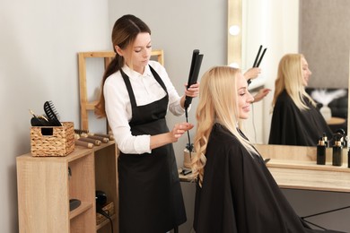 Photo of Hairdresser curling woman's hair with flat iron in salon