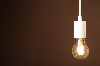 Photo of Light bulb hanging on cord against brown background, space for text