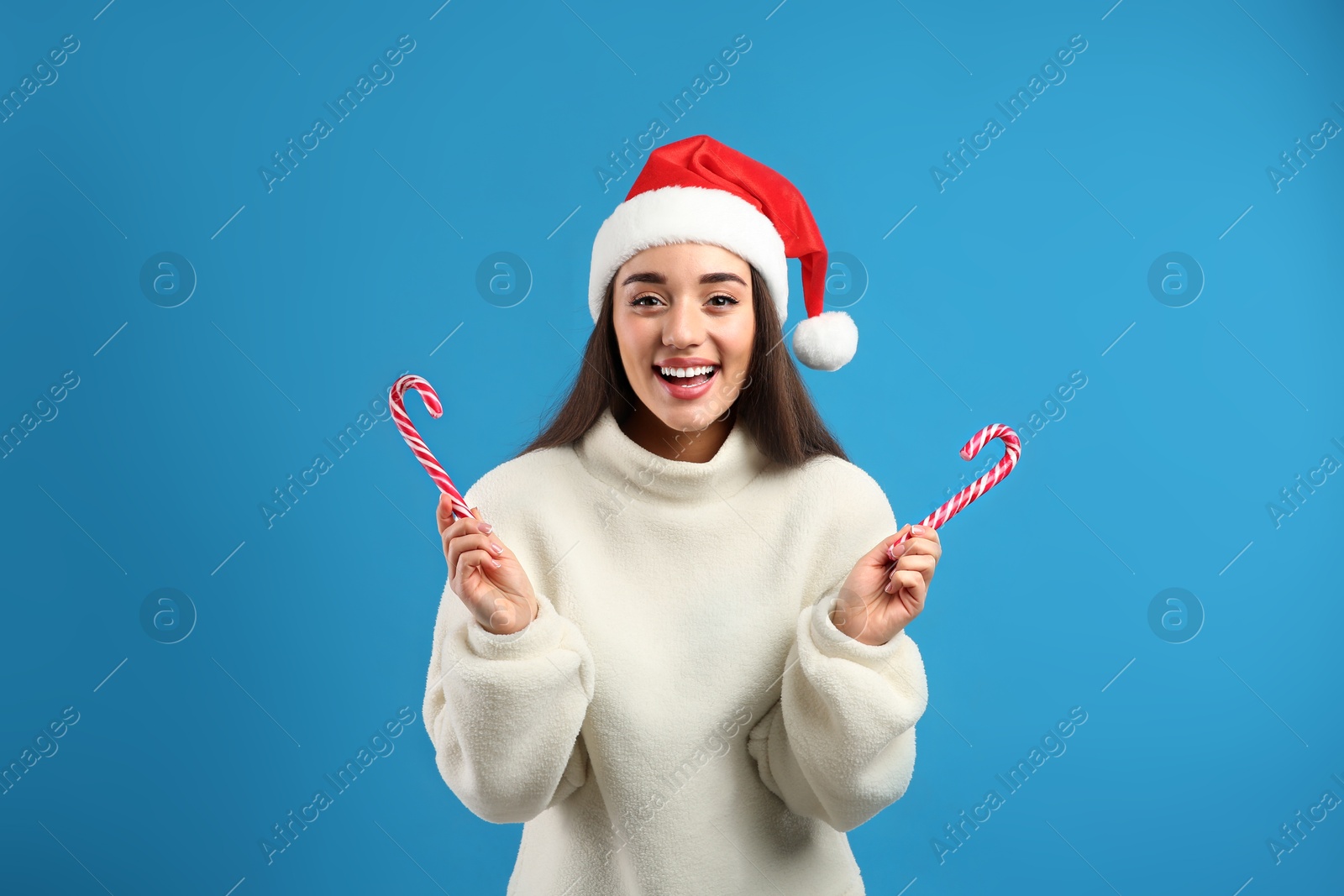 Photo of Young woman in beige sweater and Santa hat holding candy canes on blue background. Celebrating Christmas