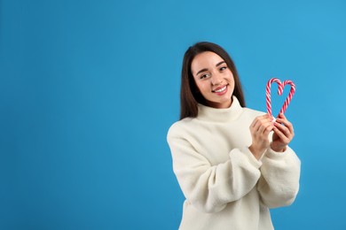 Photo of Young woman in beige sweater holding candy canes on blue background, space for text. Celebrating Christmas