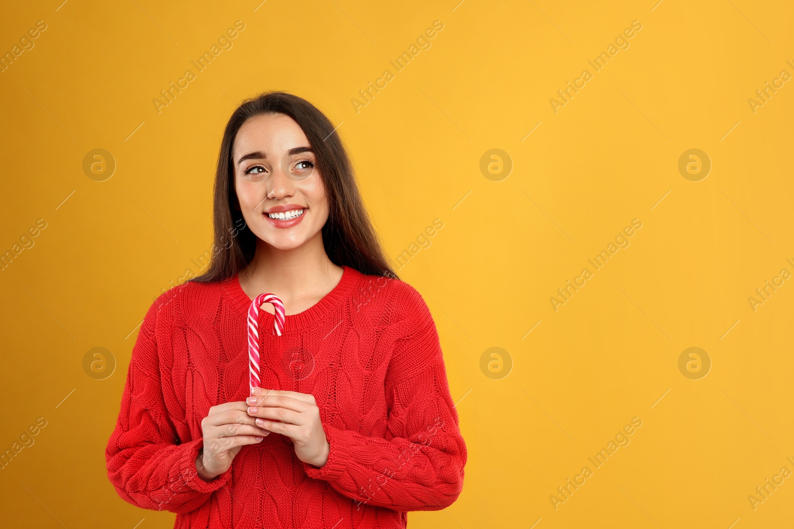 Photo of Young woman in red sweater holding candy cane on yellow background, space for text. Celebrating Christmas