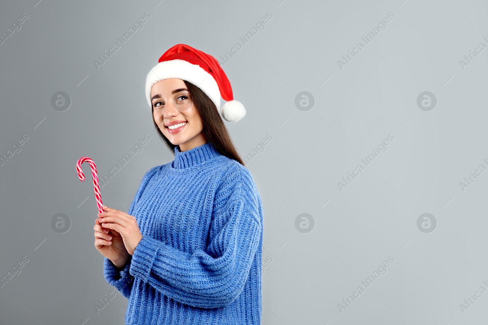 Photo of Young woman in blue sweater and Santa hat holding candy cane on grey background. Space for text