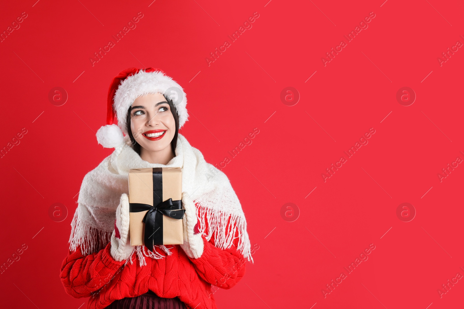 Photo of Woman in Santa hat, knitted mittens, scarf and sweater holding Christmas gift on red background, space for text