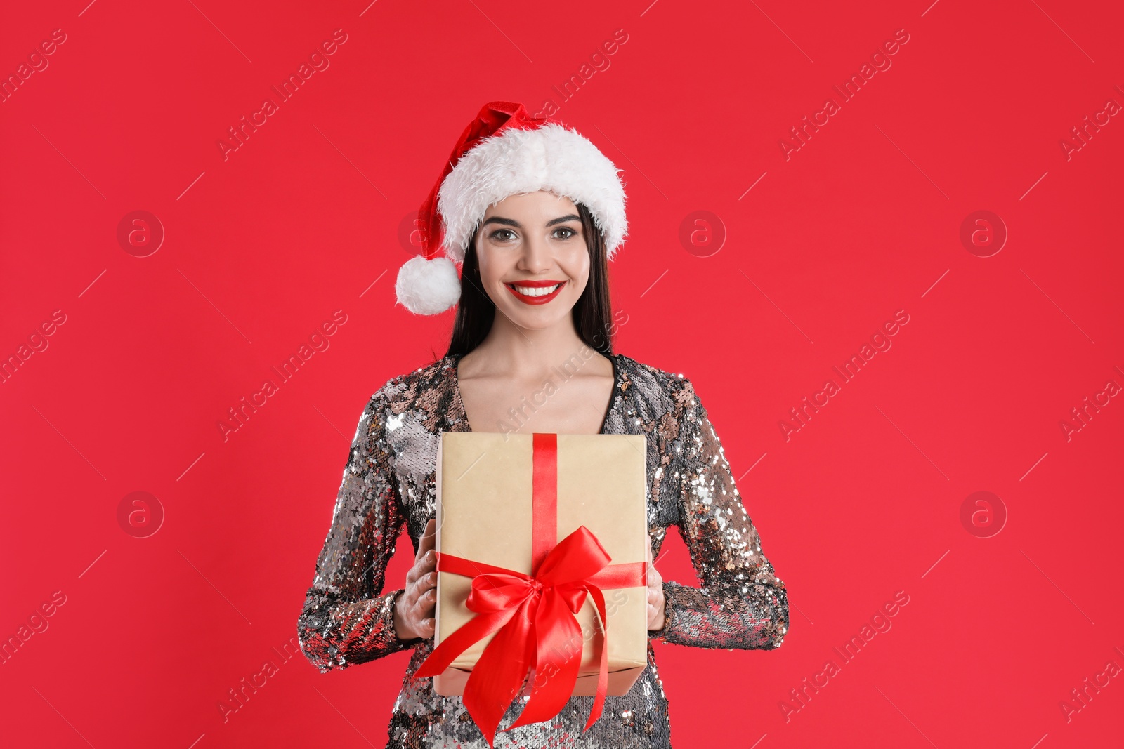 Photo of Woman in silver dress and Santa hat holding Christmas gift on red background