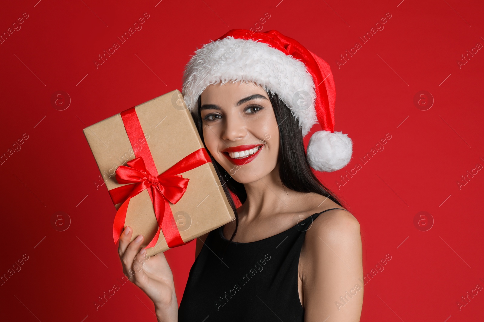 Photo of Woman in black dress and Santa hat holding Christmas gift on red background