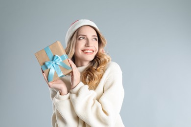 Beautiful young woman in Santa hat with Christmas present on light grey background