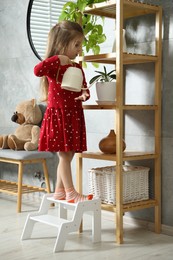 Photo of Little girl standing on step stool and watering plants on shelf indoors