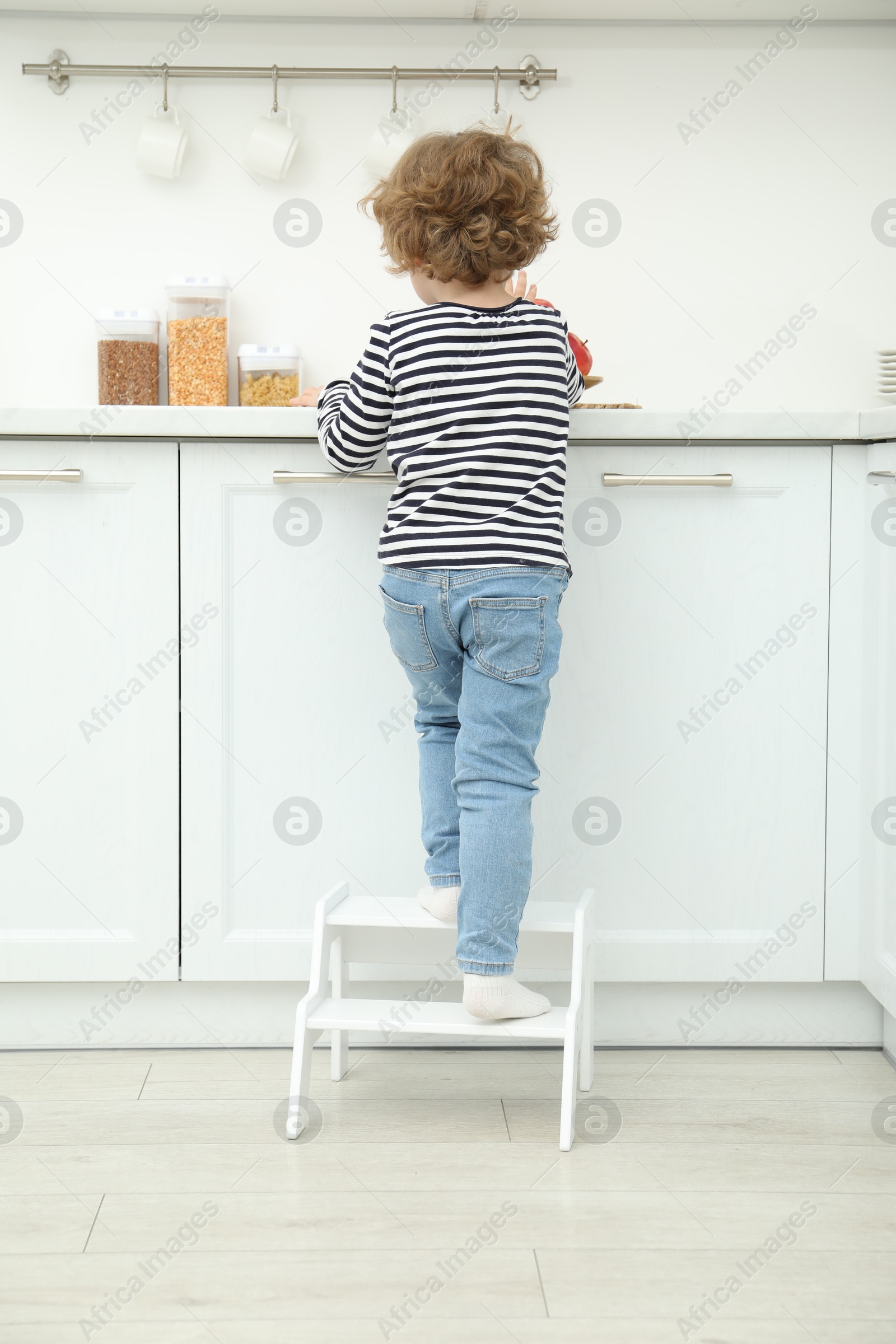 Photo of Little boy standing on step stool near countertop in kitchen, back view