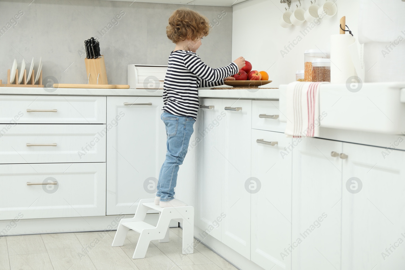 Photo of Little boy standing on step stool near countertop with fruits in kitchen