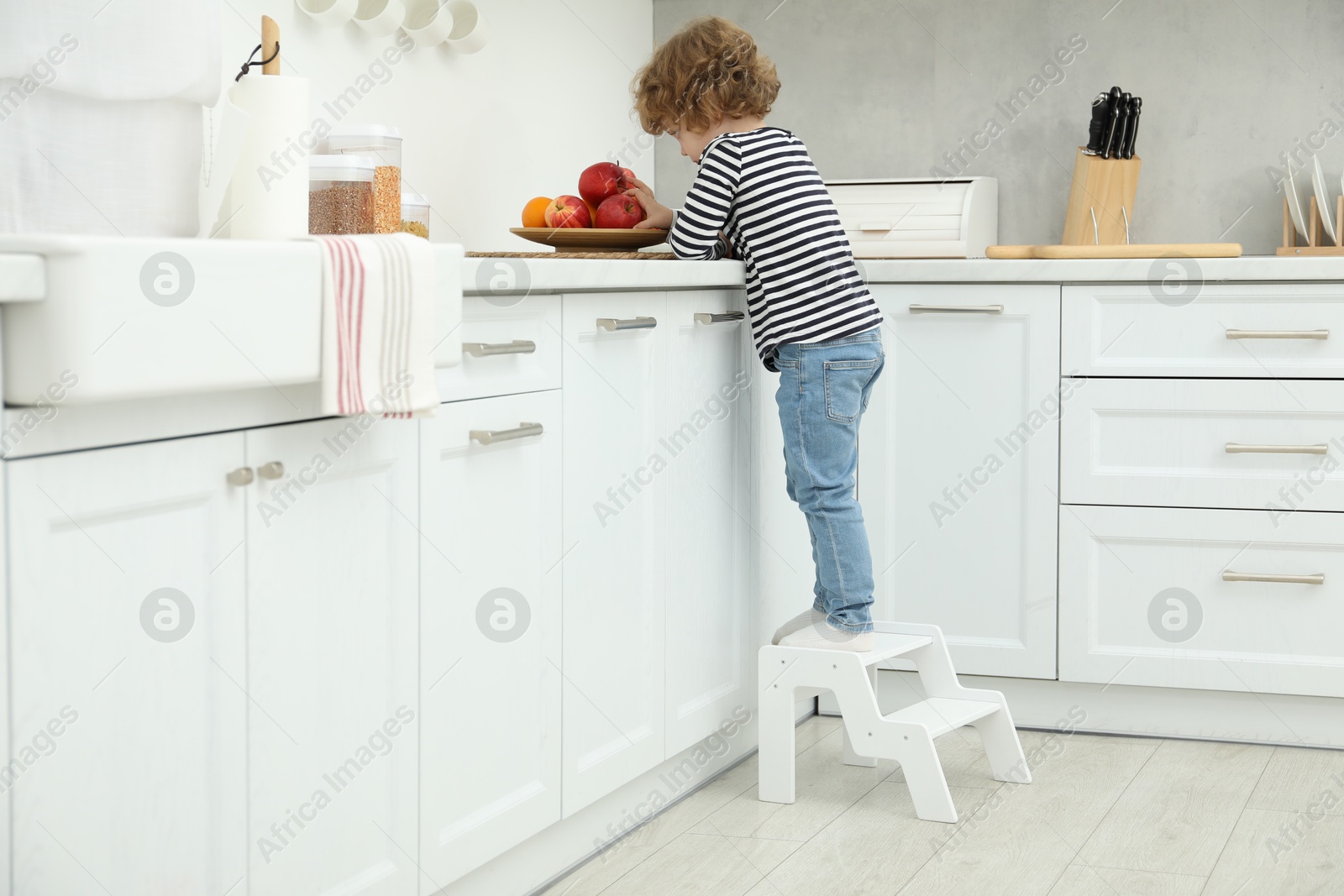 Photo of Little boy standing on step stool near countertop with fruits in kitchen