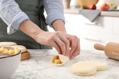 Photo of Woman making pirozhki (stuffed pastry pies) with apples at white marble table indoors, closeup