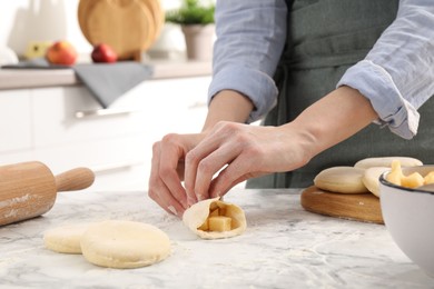 Photo of Woman making pirozhki (stuffed pastry pies) with apples at white marble table indoors, closeup