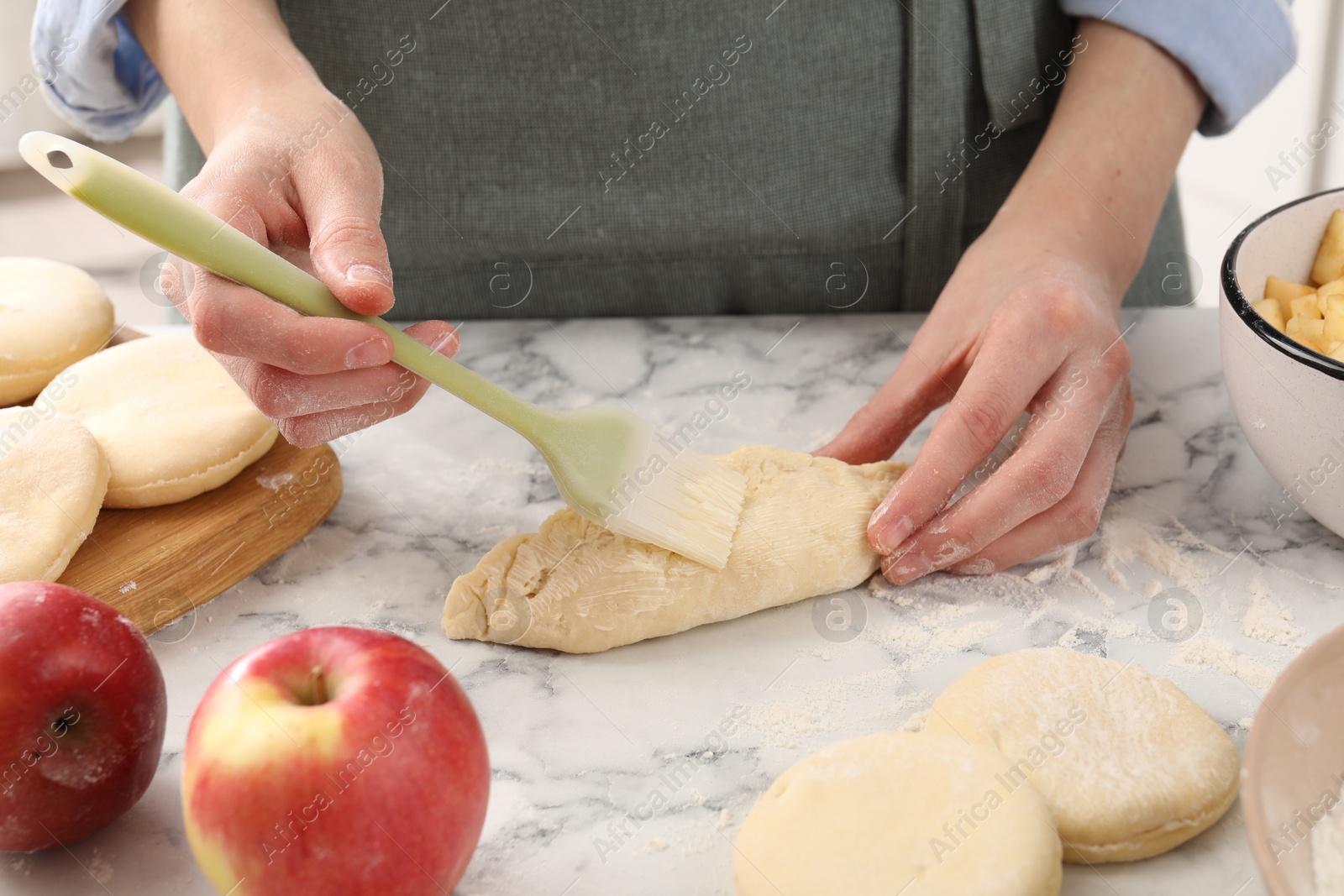 Photo of Woman brushing egg yolk onto pirozhok (stuffed pastry pie) at white marble table, closeup