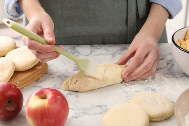 Photo of Woman brushing egg yolk onto pirozhok (stuffed pastry pie) at white marble table, closeup