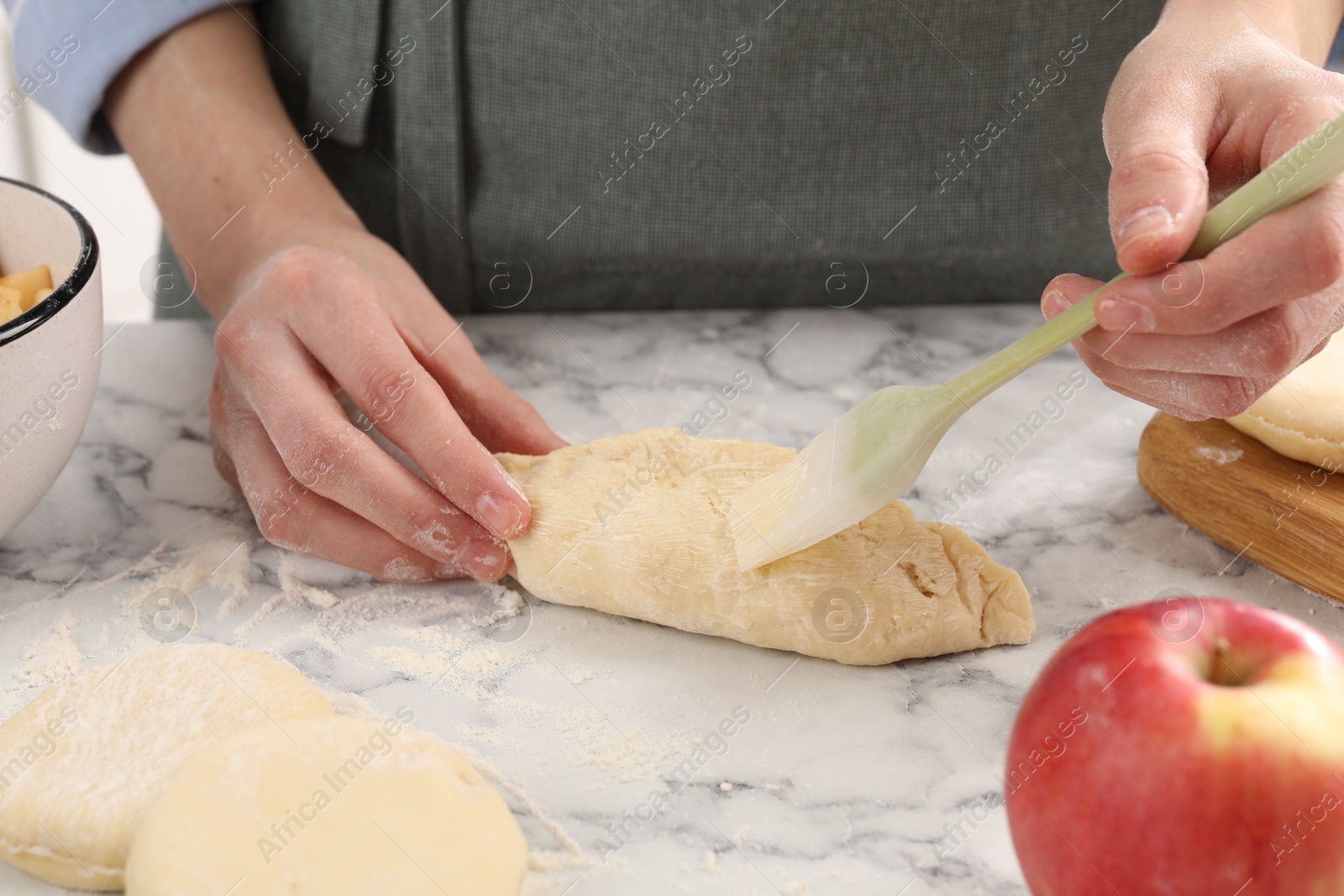 Photo of Woman brushing egg yolk onto pirozhok (stuffed pastry pie) at white marble table, closeup