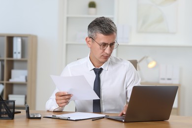 Businessman with document working on laptop at table in office