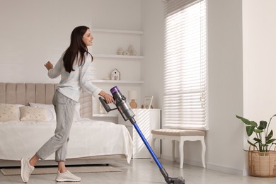 Photo of Young woman cleaning floor with cordless vacuum cleaner in bedroom