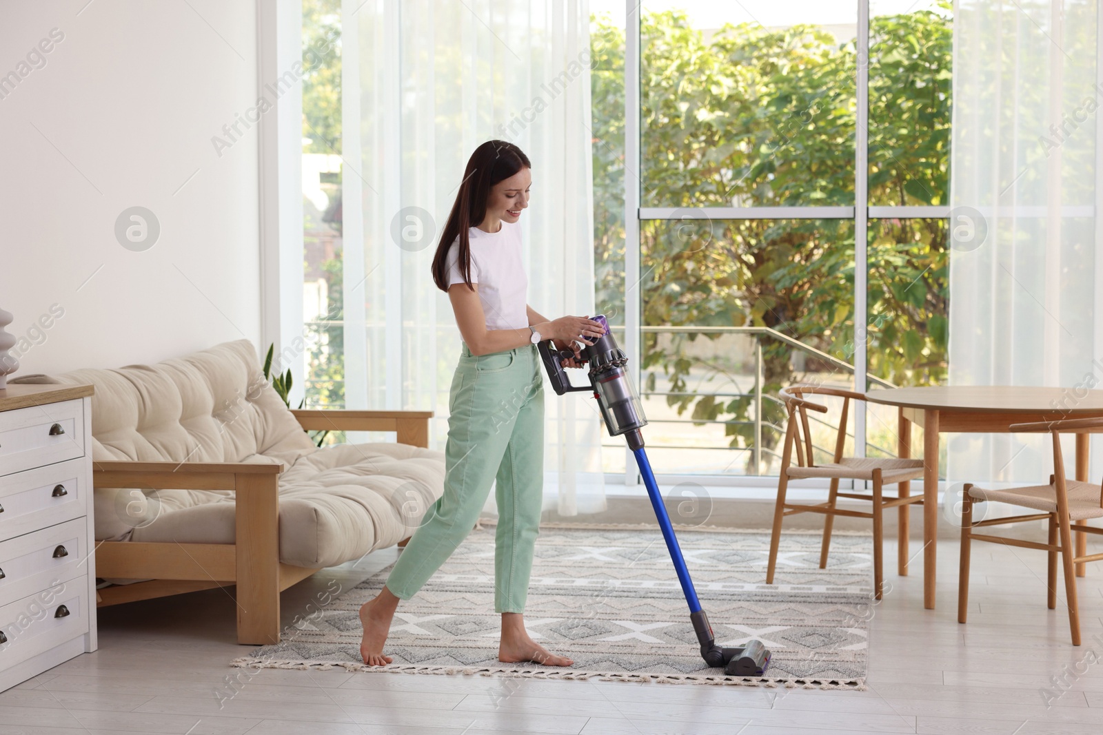 Photo of Smiling young woman cleaning floor with cordless vacuum cleaner at home