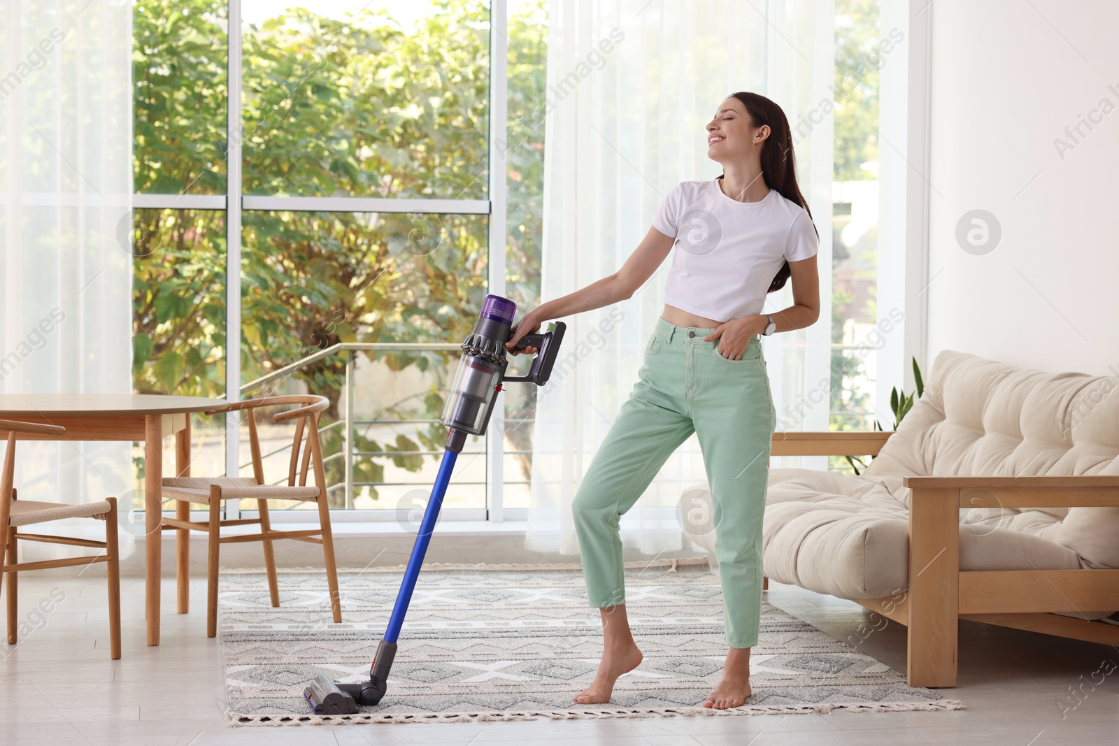 Photo of Smiling young woman cleaning floor with cordless vacuum cleaner at home