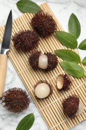 Photo of Delicious ripe rambutans, knife and green leaves on white marble table, flat lay