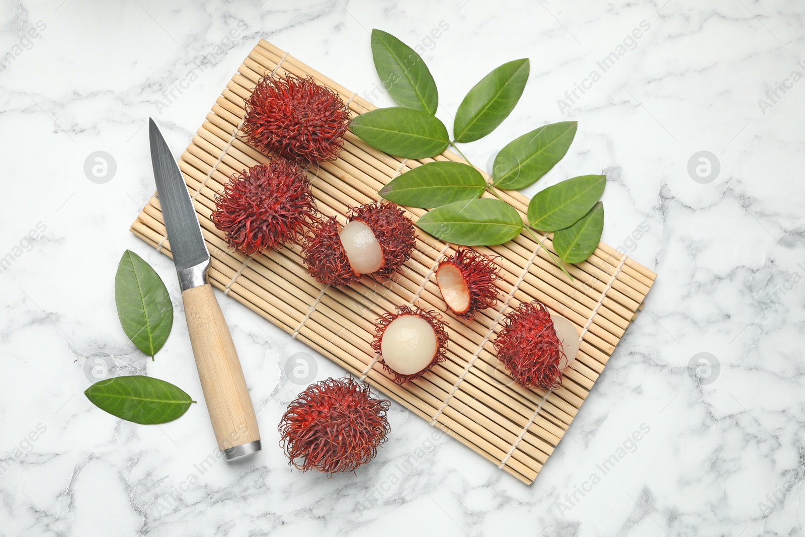 Photo of Delicious ripe rambutans, knife and green leaves on white marble table, flat lay