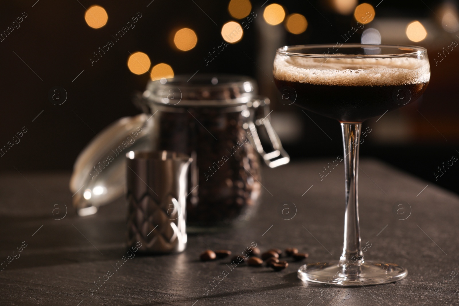 Photo of Glass with delicious espresso martini and coffee beans on dark table against blurred lights, closeup