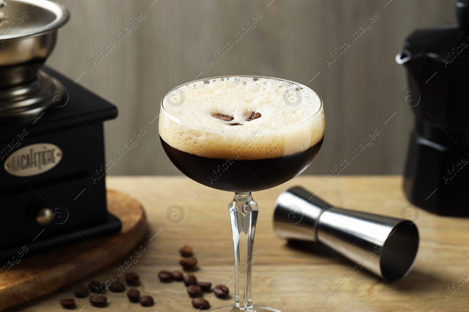 Photo of Delicious espresso martini in glass, coffee beans, manual grinder and jigger on wooden table, closeup