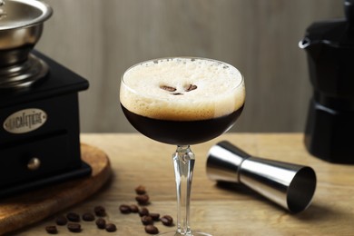 Photo of Delicious espresso martini in glass, coffee beans, manual grinder and jigger on wooden table, closeup