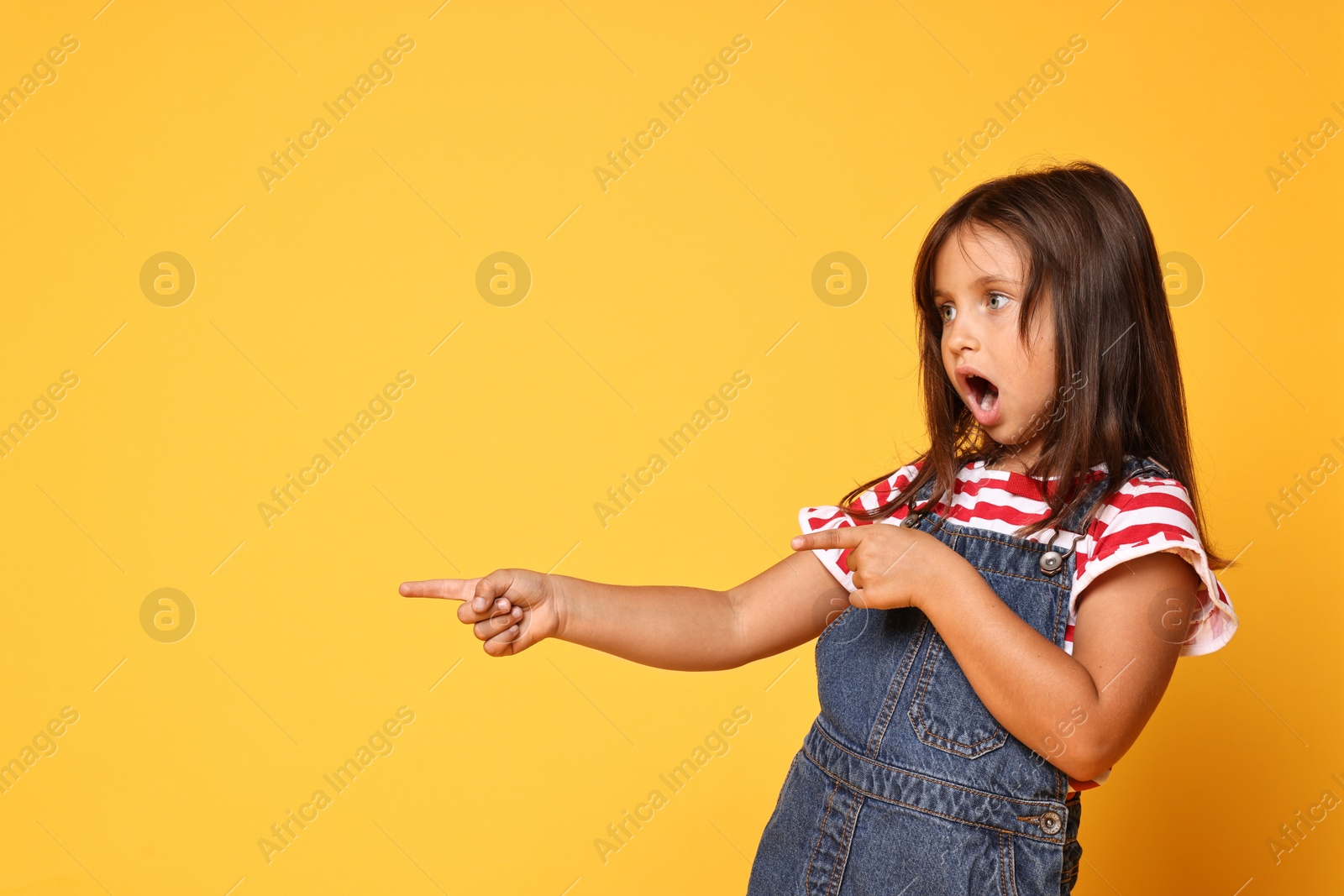 Photo of Scared little girl pointing at something on orange background. Space for text