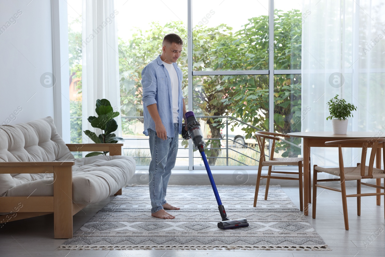 Photo of Young man cleaning rug with cordless vacuum cleaner at home