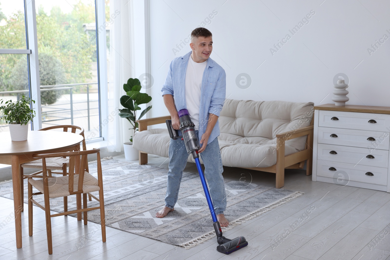 Photo of Young man cleaning floor with cordless vacuum cleaner at home