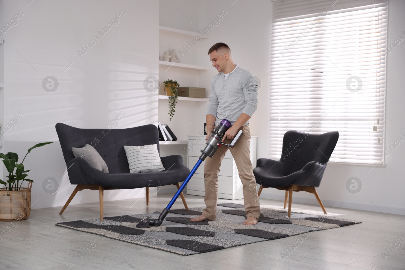 Photo of Young man cleaning rug with cordless vacuum cleaner at home