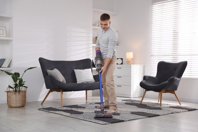 Photo of Young man cleaning rug with cordless vacuum cleaner at home